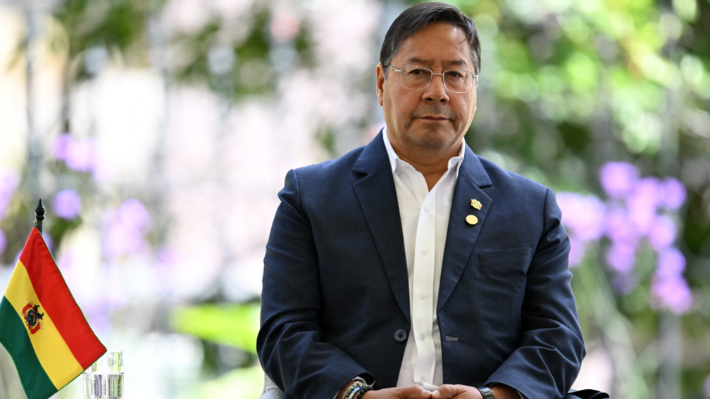 Bolivia's Luis Arce Catacora attends the Amazon Cooperation Treaty Organization (OTCA) Summit in Bogota when he was still president. He is wearing a navy suit with a white shirt and is sitting next to the national flag of Bolivia