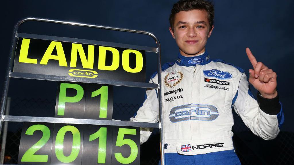 Lando Norris holds up a pit board that says 'Lando P1 2015'