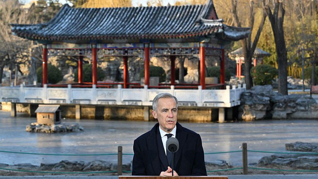 Canada's Prime Minister Mark Carney, wearing a suit and tie, speaks into a mike. Behind him is a structure with a traditional Chinese roofing style, with upturned eaves