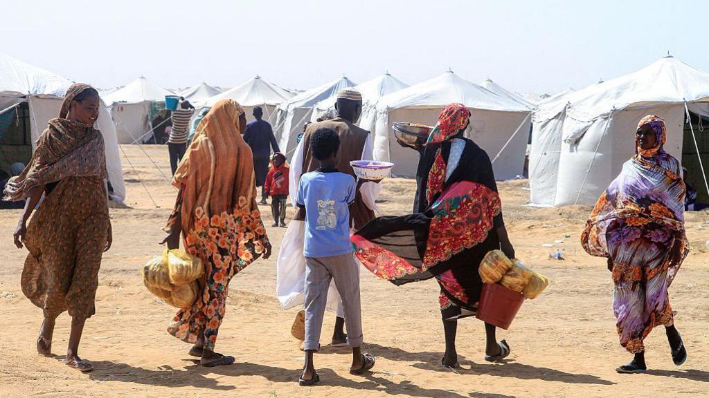 Four women, a boy and a man walking on sandy ground carrying food. In the background are white aid tents.