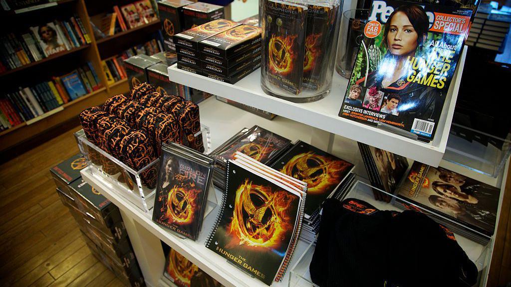 A shelf in a bookshop loaded with Hunger Games books and stationery bearing the series logo of a golden bird inside a burning ring, and magazines bearing portraits of Jennifer Lawrence.