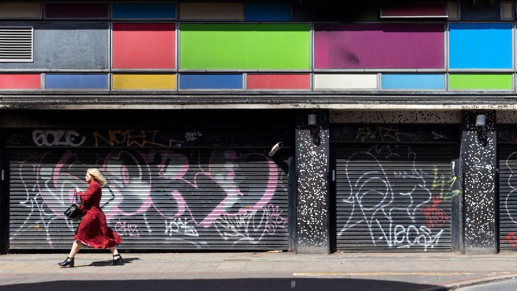 A woman walks past two shops with the shutters closed.