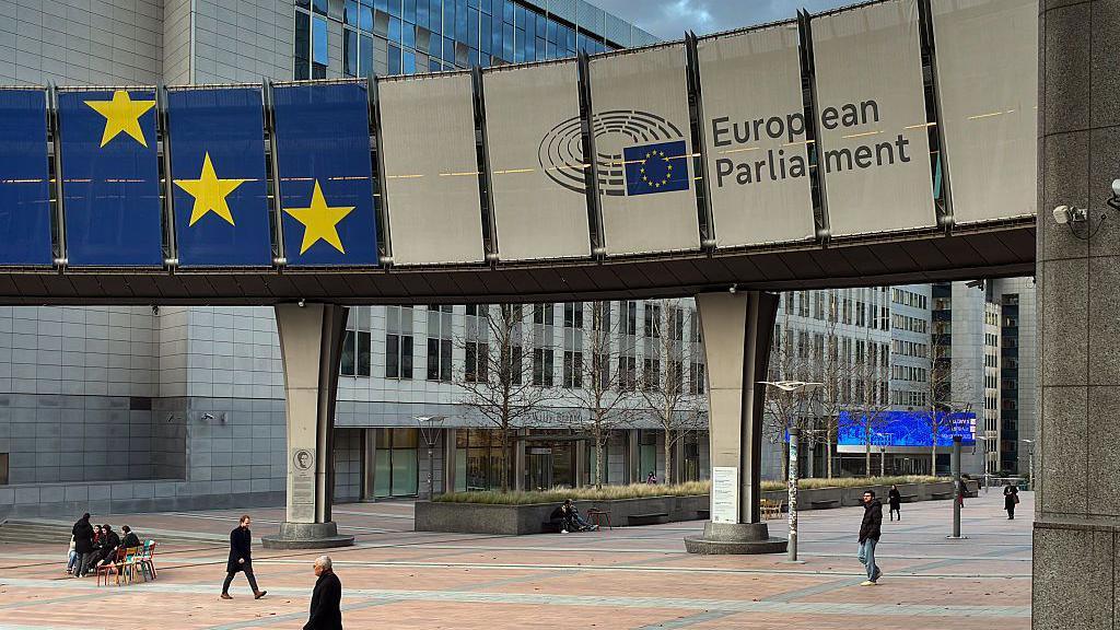 An exterior view of the Altiero Spinelli Building, part of the legislative complex of the European Parliament, shows a banner sign with the blue-and-yellow EU flag and the institution's name displayed on an elevated walkway.