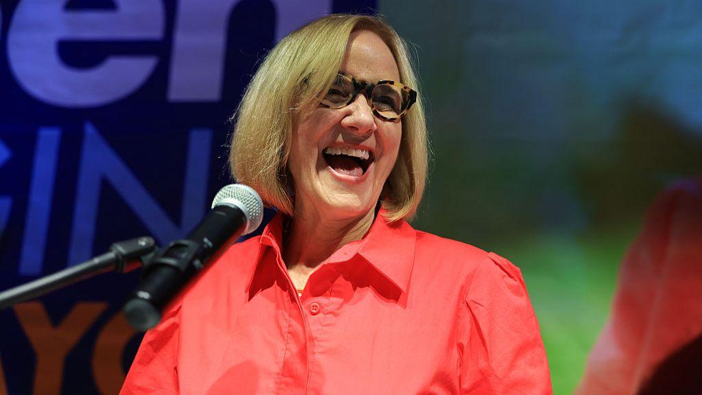 Eileen Higgins, wearing a bright pink collared shirt and tortoise shell glasses, smiles in front of a microphone after winning the Miami mayoral race.
