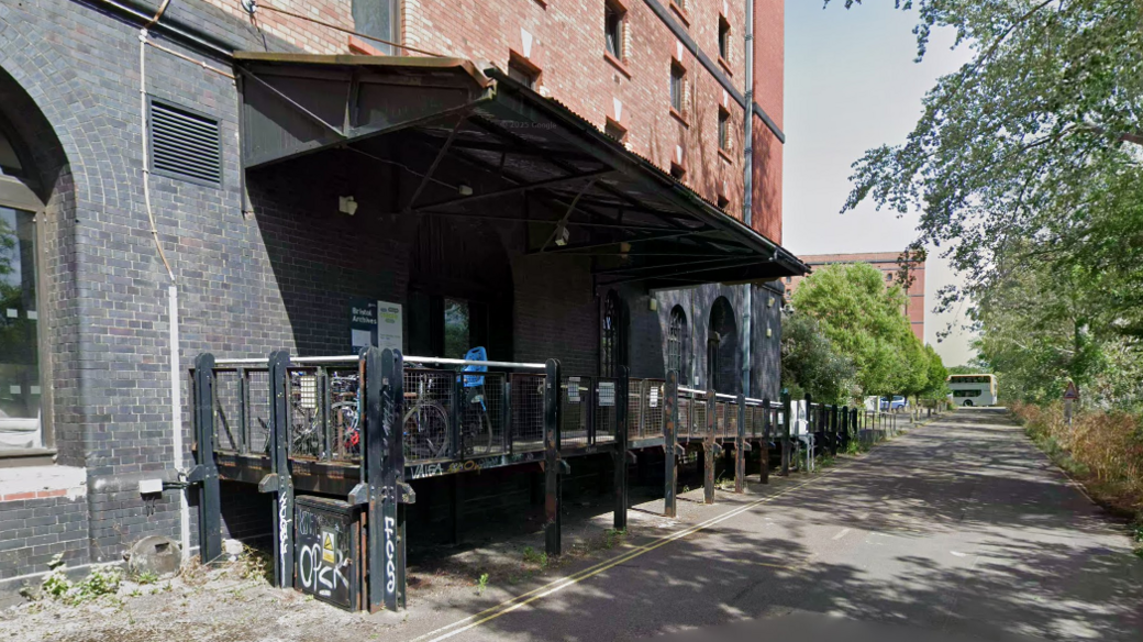 A large, black and red brick building with a black metal ramp leading to it. There is a road outside the building flanked by trees. Bikes are chained up outside the building.