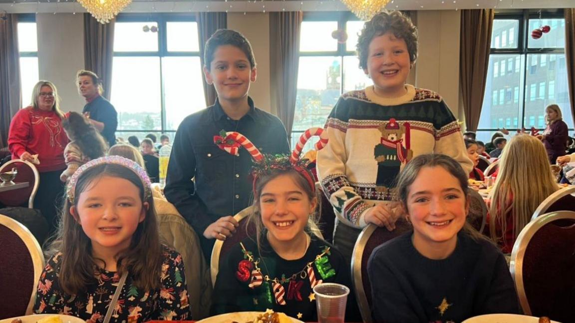 Five children smiling at the camera. At the back there are two boys and in the front three girls. All of them are wearing Christmas themed outfits. Behind them are windows.