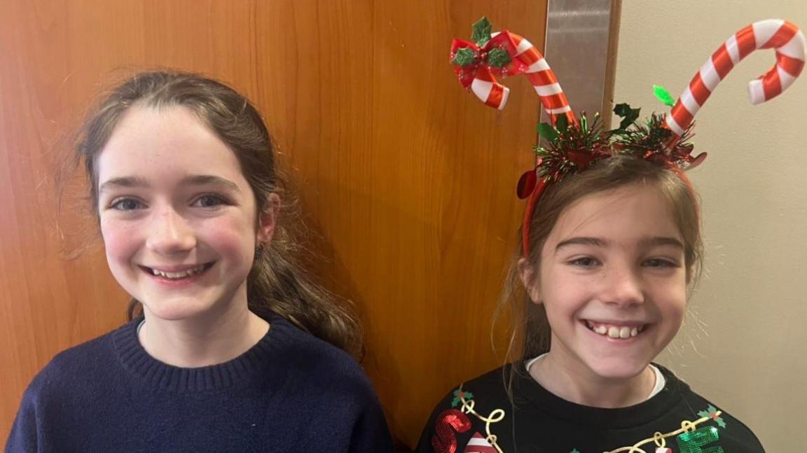 Two girls smiling at the camera. One of the girls is wearing a candy cane headband. Behind them is an oak door.
