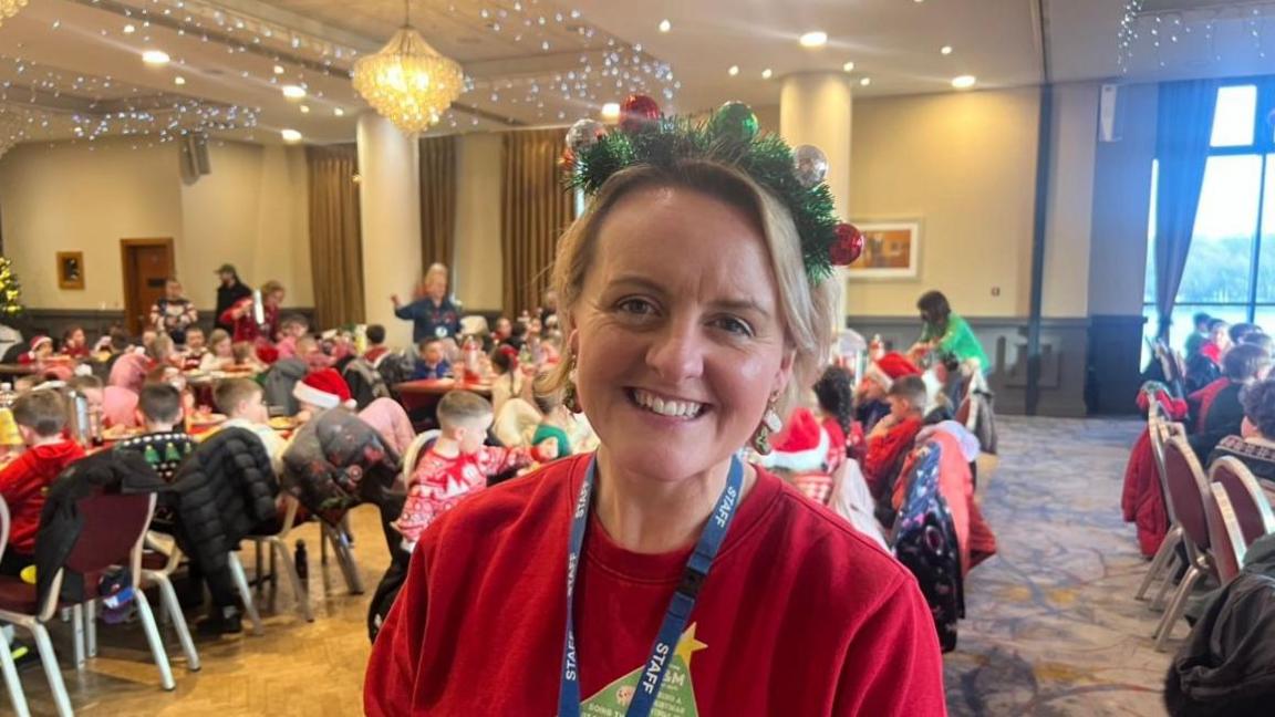 Women wearing a red jumper and a Christmas hat on. Behind her are school children and Christmas lights.