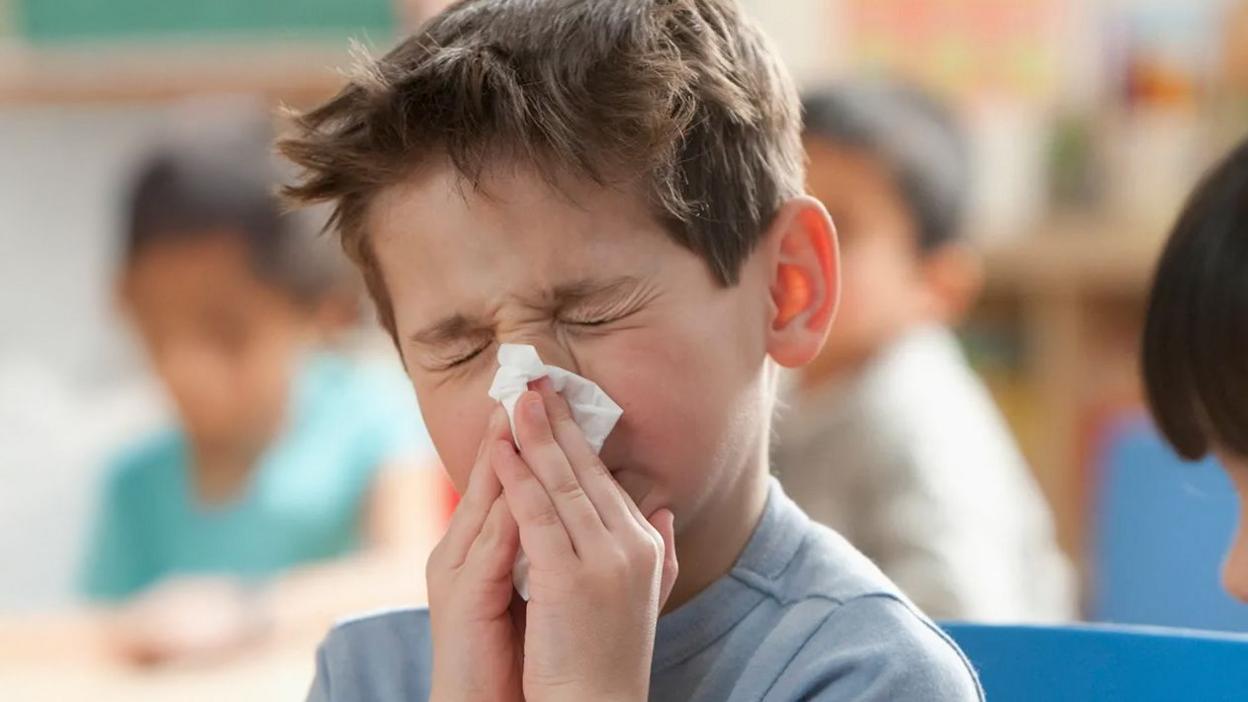 Child sitting indoors with hands near mouth, wearing a light blue top, in a classroom setting with other children and colorful background elements.