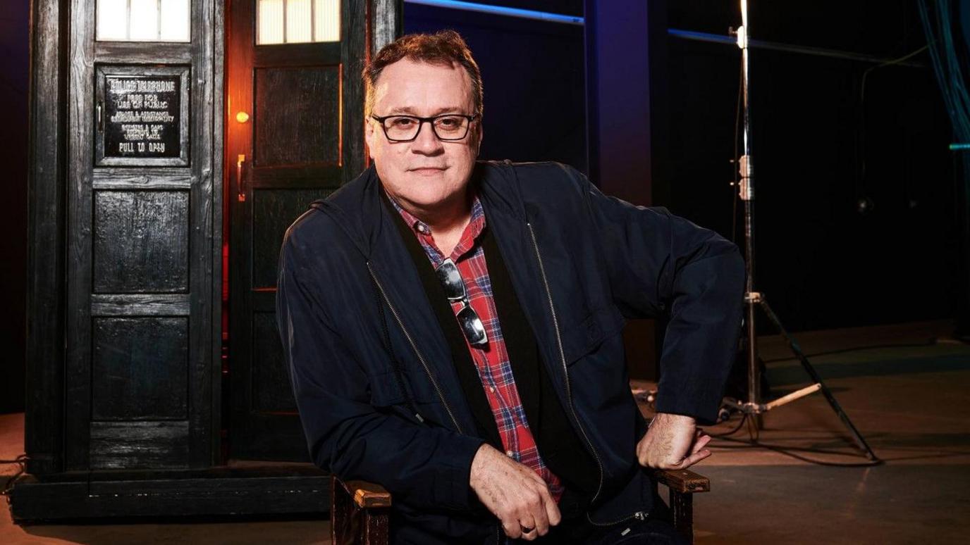 writer Russell T Davies is wearing a red and blue checked shirt with a black jacket. It's a professional photograph taken on the set of Dr Who. You can see the Tardis in the background. He has dark hair and wears black glasses. He is resting one elbow on the arm of his seat and his knuckles are facing down on the other armrest.
