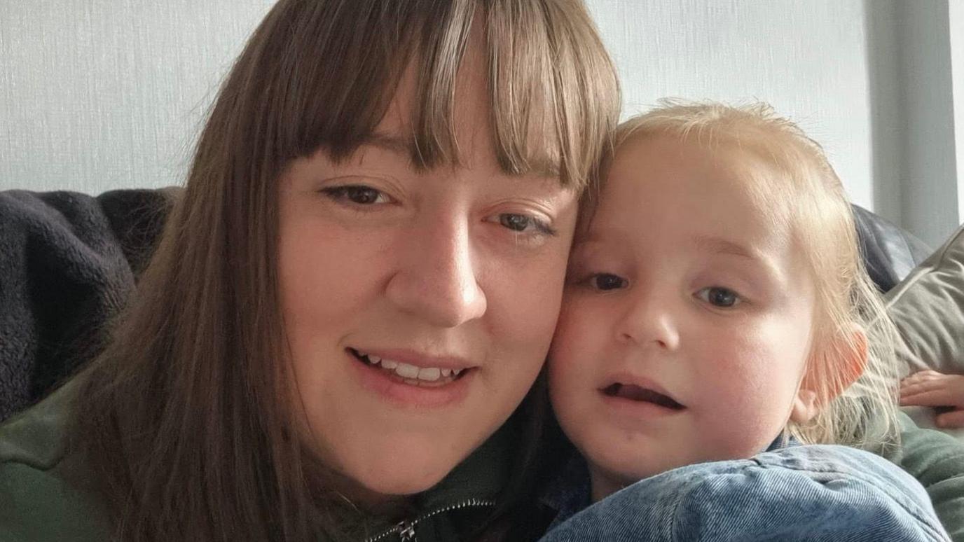 Dorothy Murray sitting on a sofa holding her young daughter Sienna. Ms Murray has long, straight brown hair with a fringe and is wearing an olive green zipped jacket. Sienna has fair hair tied back. They are both looking into the lens.
