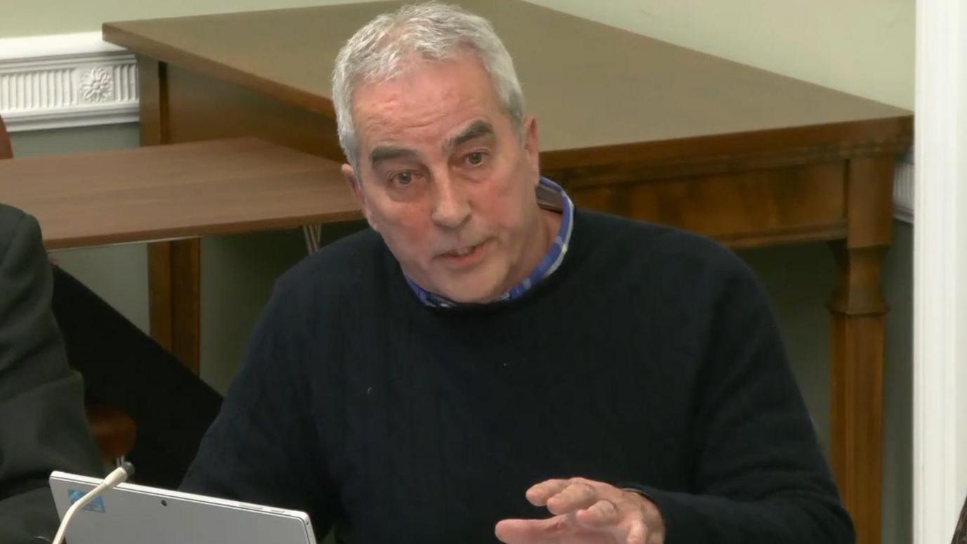 Pat Sheehan. He is wearing a black jumper with a blue shirt. He has grey hair.