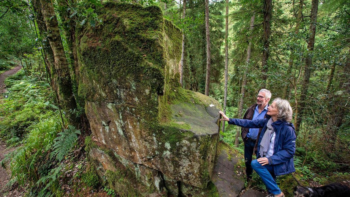 Older man and women, both with grey hair, gaze at a two metre high rock shaped like a chair. The woman is rested the fingertips of her outstretched arm on the slippery, mossy stone.