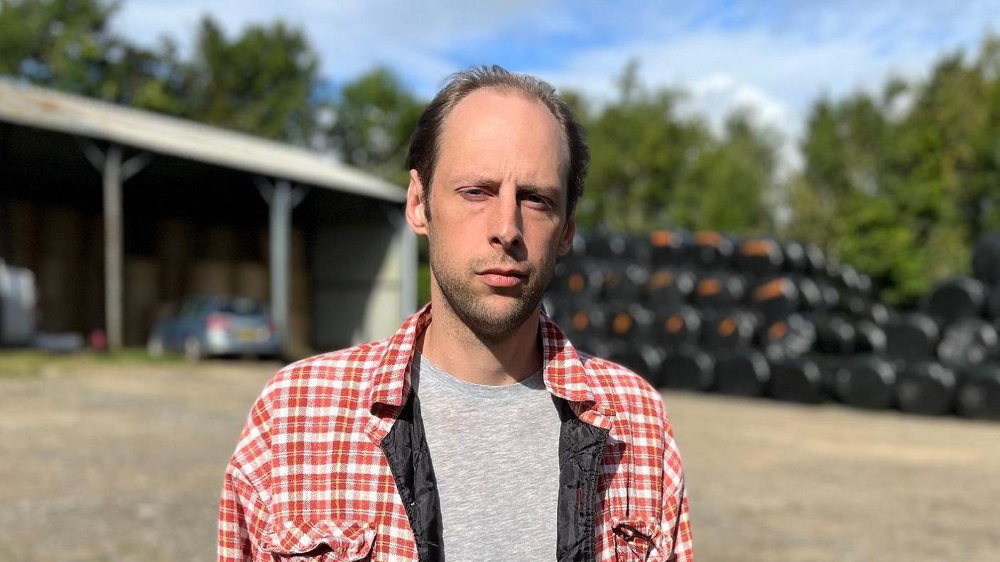 Merlin Lewis , who has brown-hair and is wearing a red checked shirt and grey T-shirt, looks at the camera. There are farm sheds and bales of hay in the background