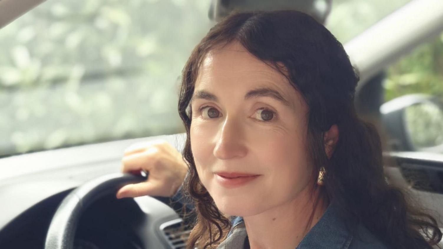 A woman with brown wavy hair sat in a car smiling.
