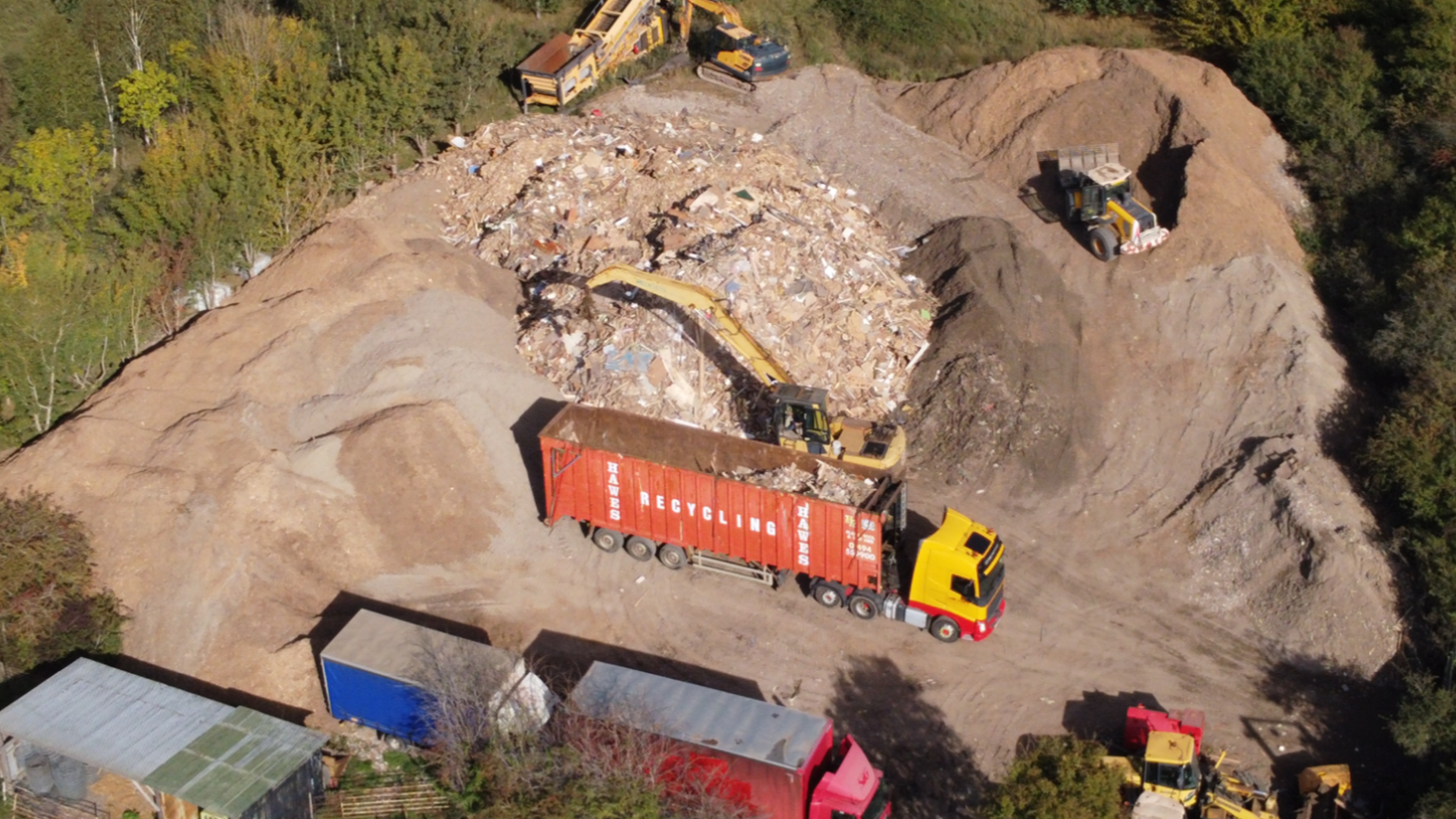 An aerial view of large piles of earth and mixed debris. Lorries are pictured moving rubble with several other excavator-type vehicles around the area, all surrounded by trees.