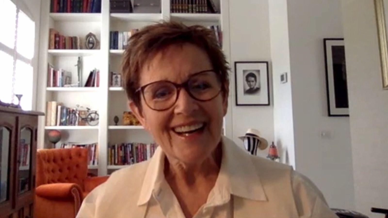A woman with short hair and glasses smiles into the camera, with an orange chair and a floor-to-ceiling bookcase behind her