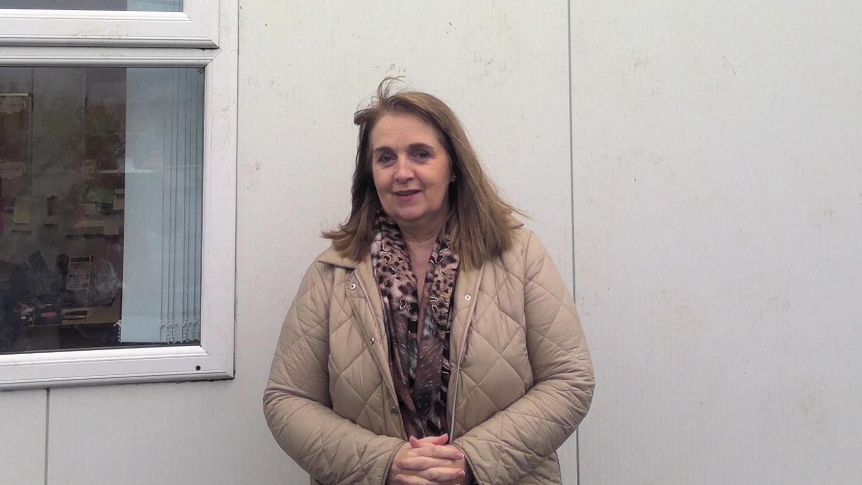 The wind has caught a a few locks of Gemma's hair, lifting part of her fringe into the air. She is wearing an animal print scarf an a beige jacket, the padding of which is separated into diamond shapes.