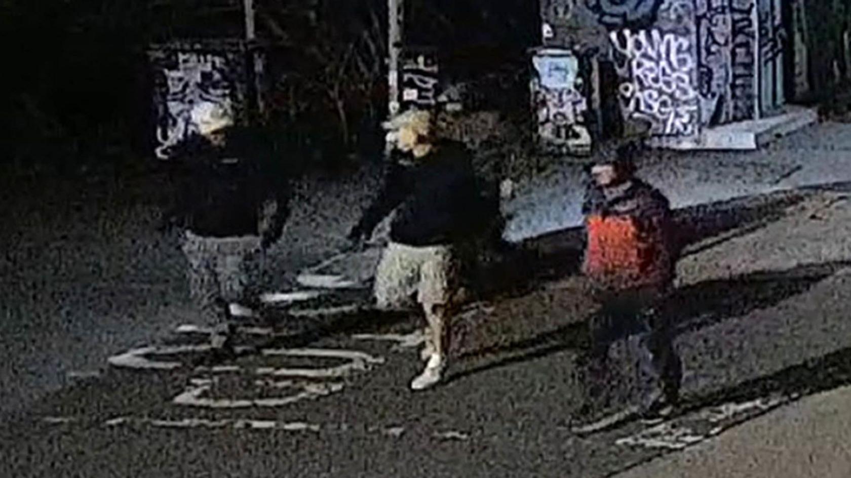 A blurry CCTV image of four men wearing jackets and baseball caps in a street at night time.