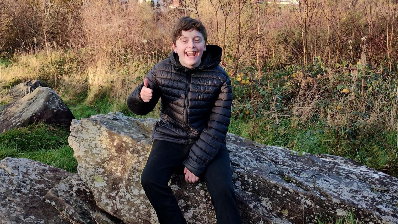 13-year-old Kyle sitting on a rock overlooking his hometown, Pontypridd. He has short black hair and is wearing a black puffer coat. He is smiling and making a thumbs up gesture.