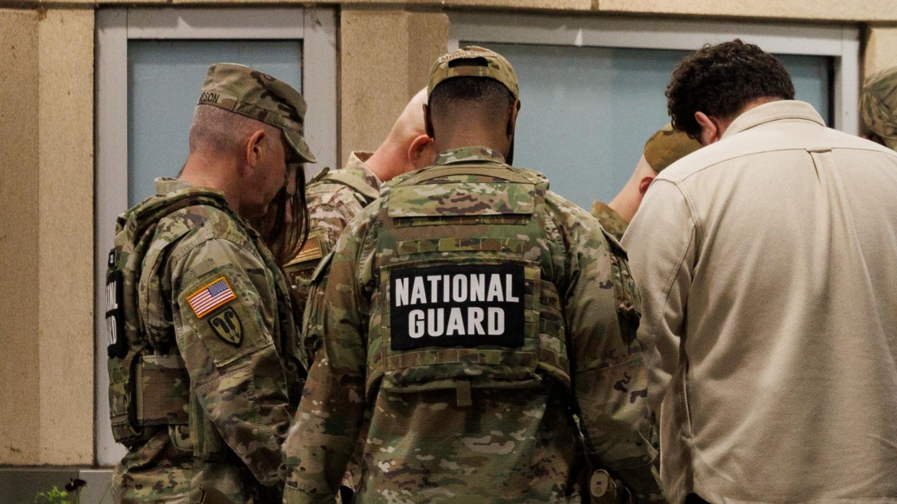 A photograph of a number of National Guard members standing together. At least 5 men can be seen in the image - one of whom appears to be in civillian clothing wearing a cream-coloured shirt. The rest of the men are in uniform and the words 'National Guard' can be seen on the back of their uniform.