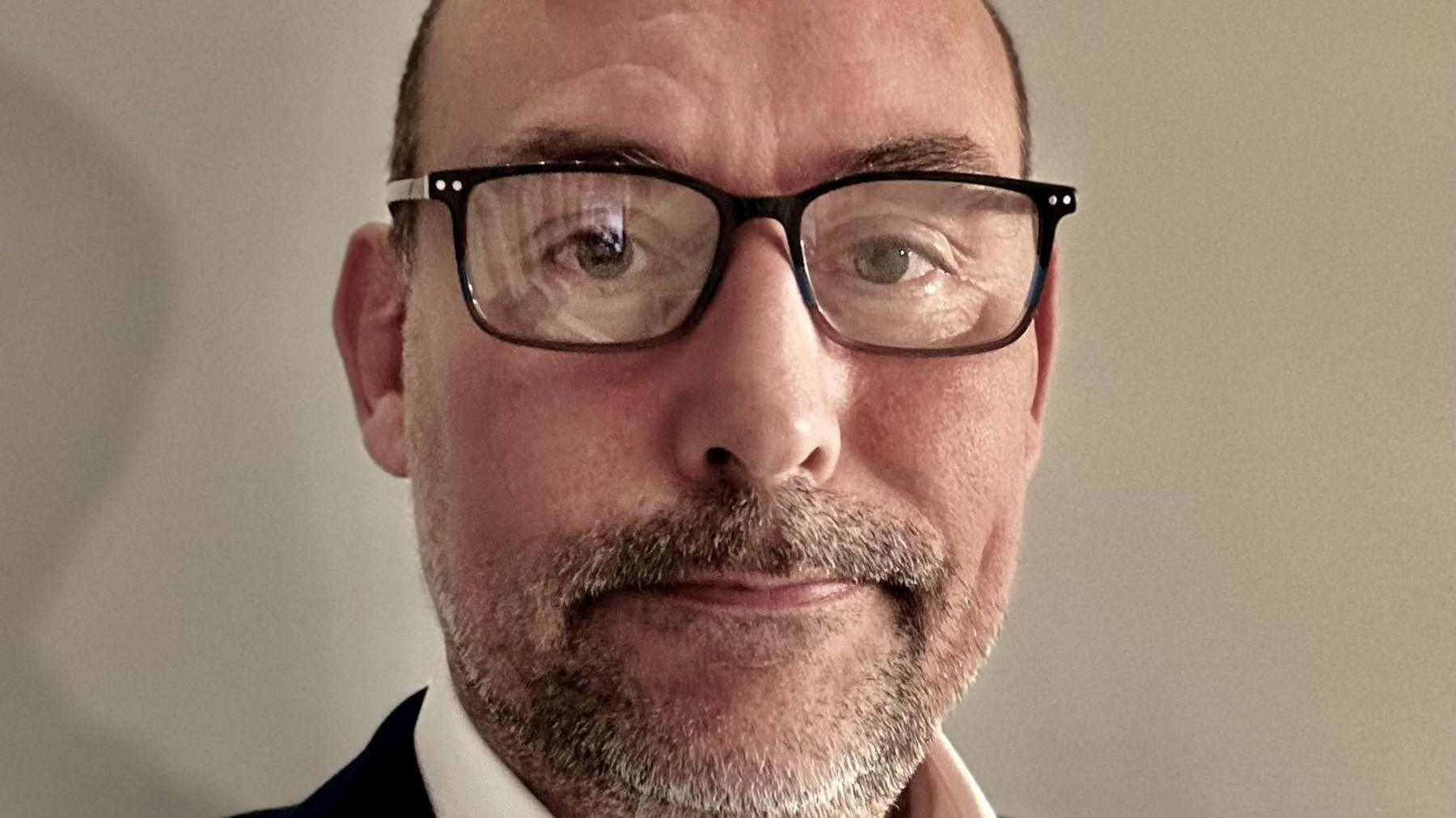 A closeup of Stephen who has very short cropped brown hair, which is receding from the forehead, and a thin beard. He is wearing square black-framed specs on his face and a dark suit.