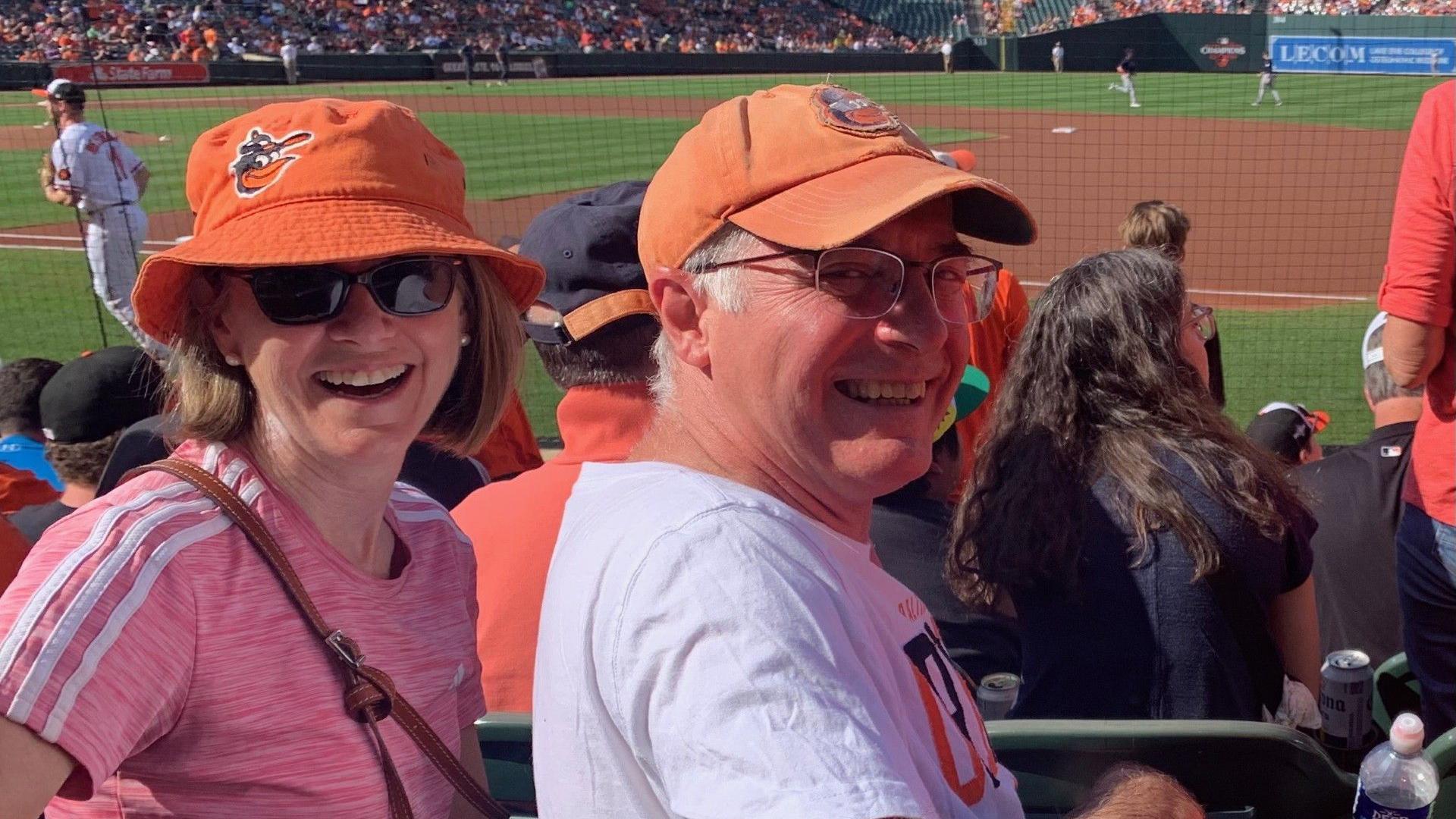 Kristy Gammon and her husband at a Baltimore Orioles game. They are both wearing orange Orioles hats and smiling. Behind them is a baseball pitch and some of the crowd.