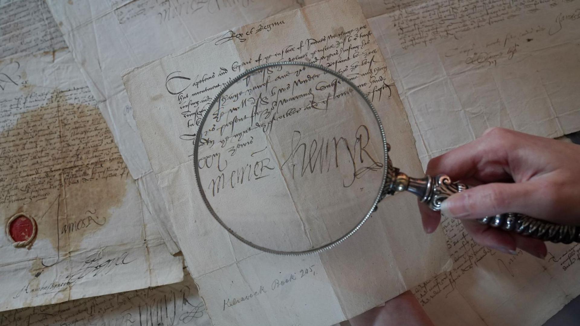 A woman holds a magnifying glass over an old letter showing the signatures of Mary, Queen of Scots and her husband Lord Darnley. There are other manuscripts laid out below the letter.