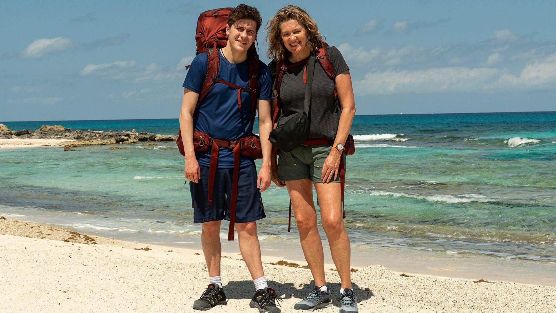 Dylan Llewellyn and mum Jackie smiling with their backpacks on whilst stood on a white sandy beach in front of the sea.