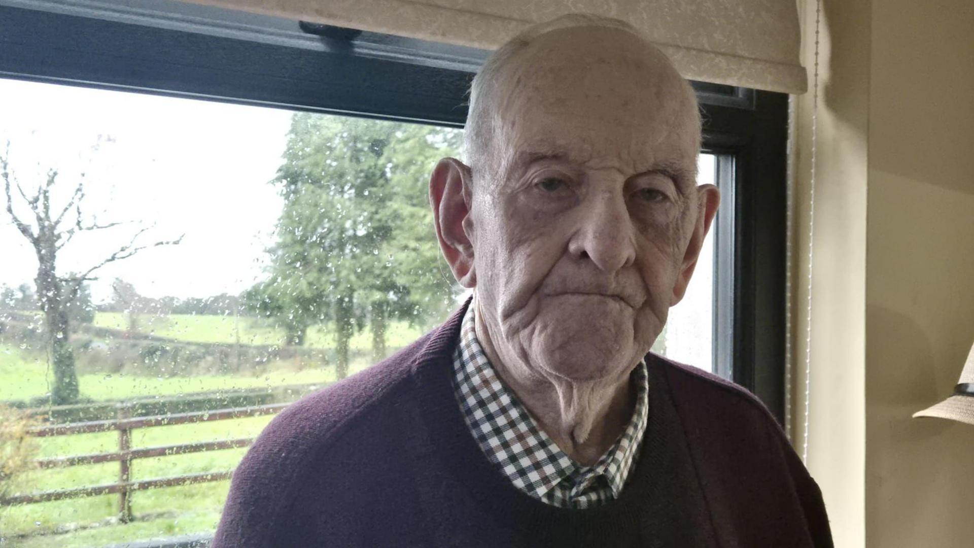 Mick's heavily receded hair is grey, and his lips are rolled inwards. He is wearing a burgundy jumper over a chequered shirt. Behind him dull light is coming through a rain-splashed window.