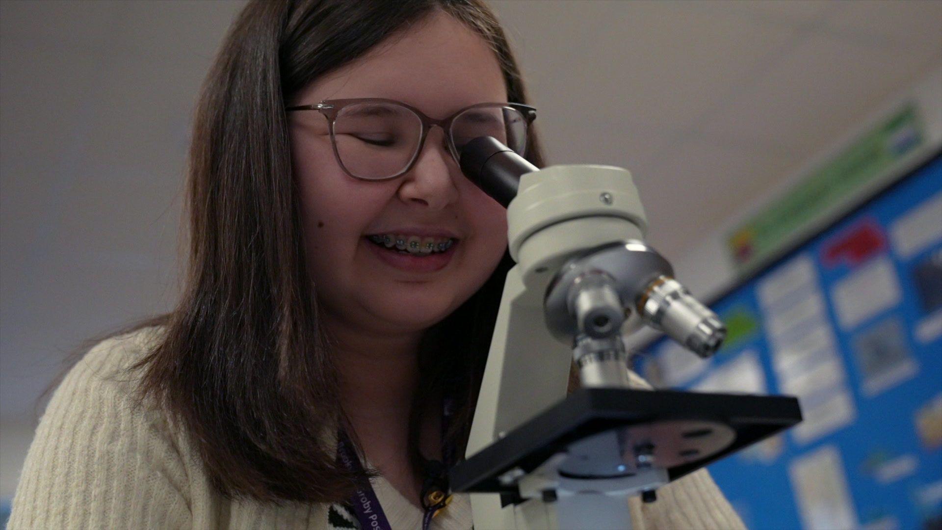 Girl with long brown hair and wearing glasses stairs down a microscope in a biology class.