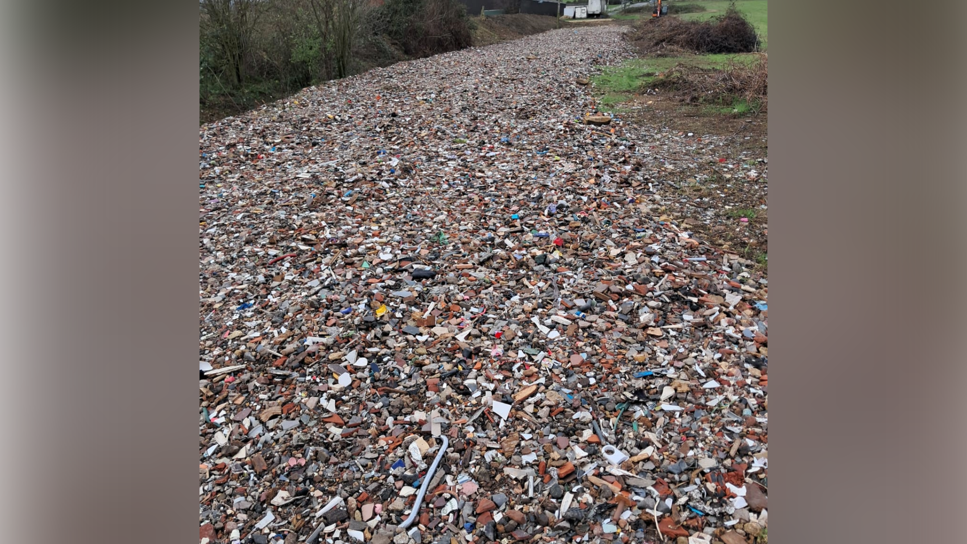 A field that is completely covered in rubble and mixed waste, including broken bricks, stones and bits of plastic. Bushes and fields line the sides.
