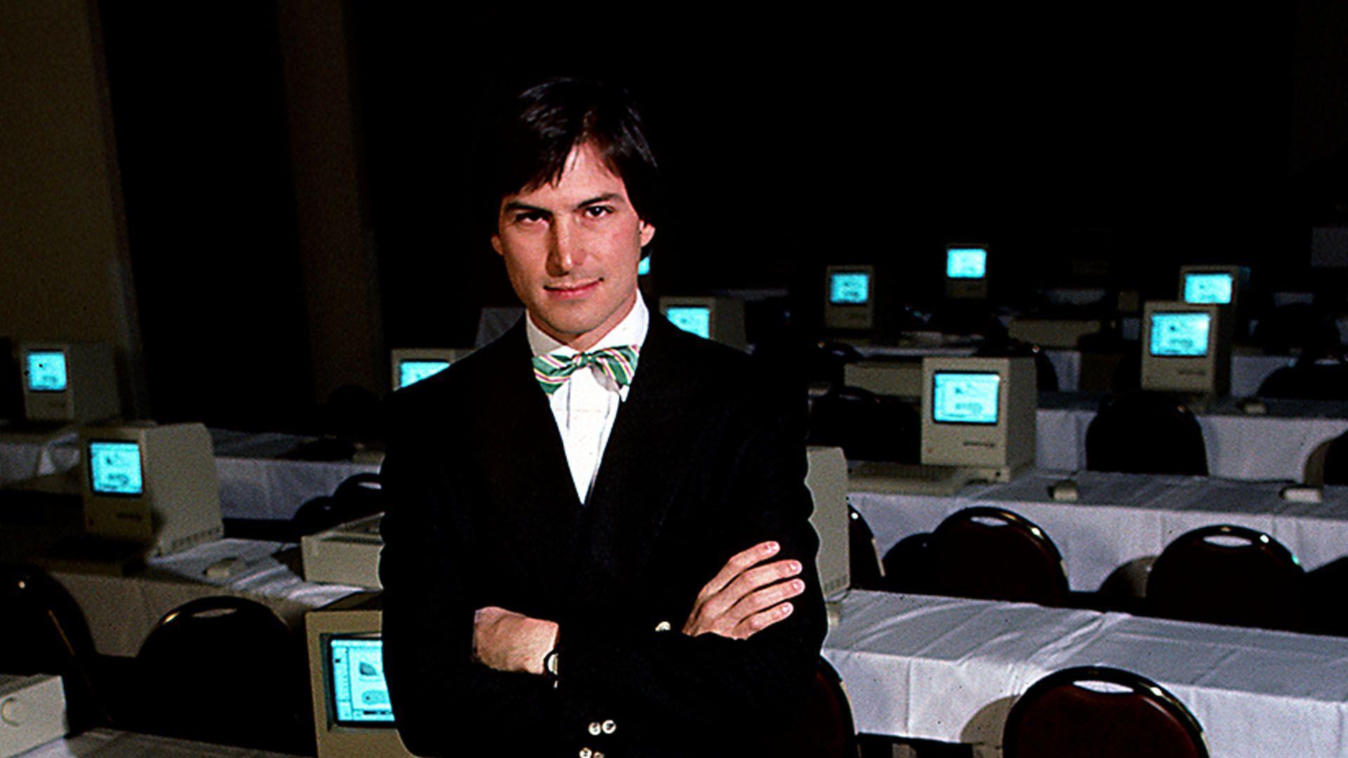 A man in a dark suit and green and yellow striped bow tie. He stands in front of a bank of computers with his arms folded.