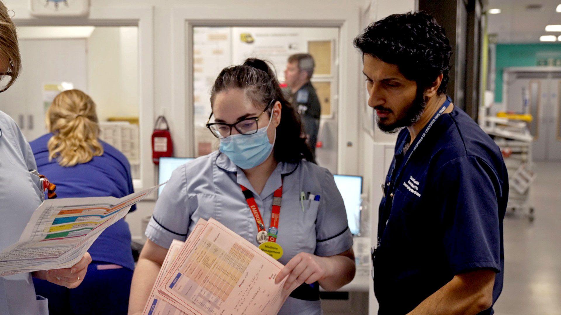 Consultant Saad Jawaid on the right is wearing blue hospital scrubs. He is speaking to female members of the clinical team, also in uniform, who are holding documents. They are in the emergency unit the the Royal Leicester Infirmary.