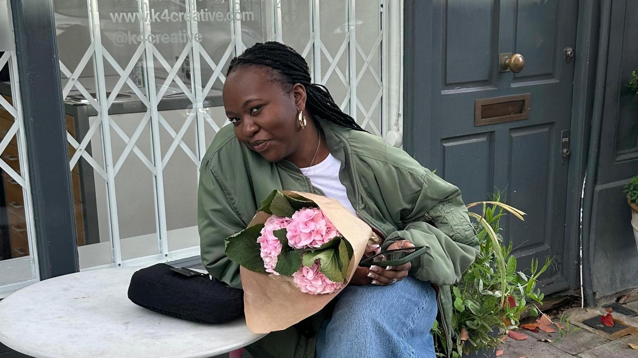 Young girl sits at a small table in front of a cafe holding pink flowers and smiling.