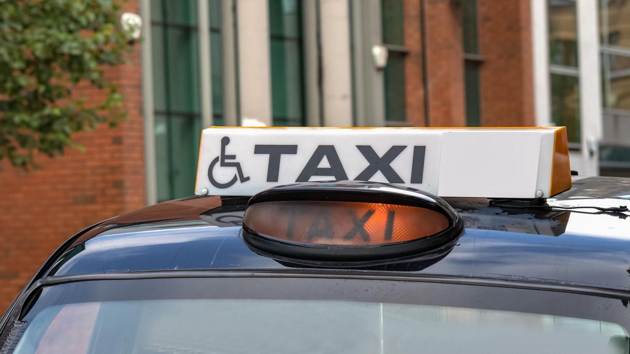 A black taxi cab with a white sign on top which reads TAXI and has a disability sign. Behind it can be seen a red brick building.