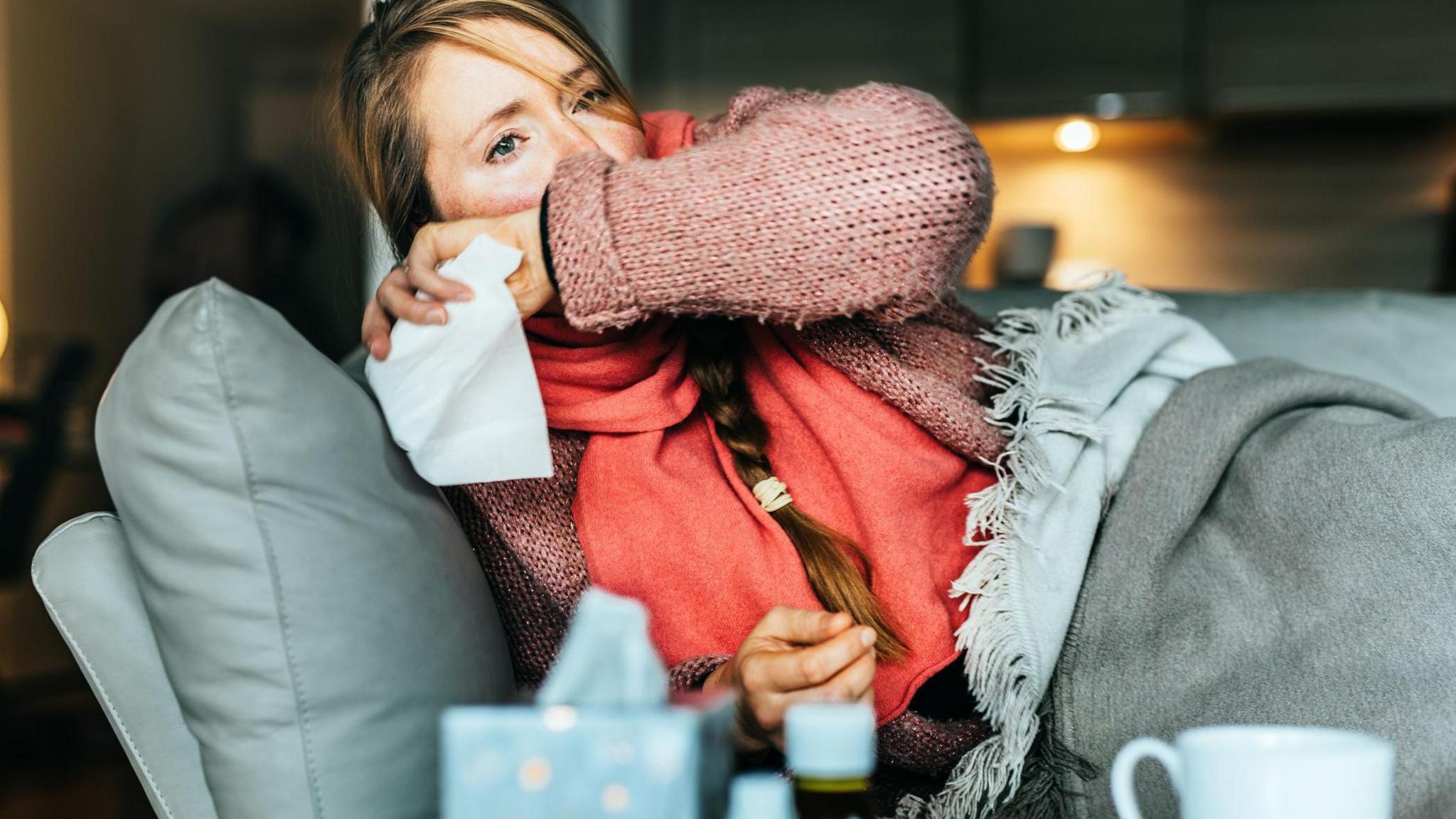 A woman with flushed cheeks lies on a couch covered in blankets. She covers her mouth, apparently coughing, and has a tissue in her hand. She has long blonde hair in a plait and is wearing a pink jumper and a coral scarf. In the foreground of the shot is a box of tissues, a mug and two medicine bottles.