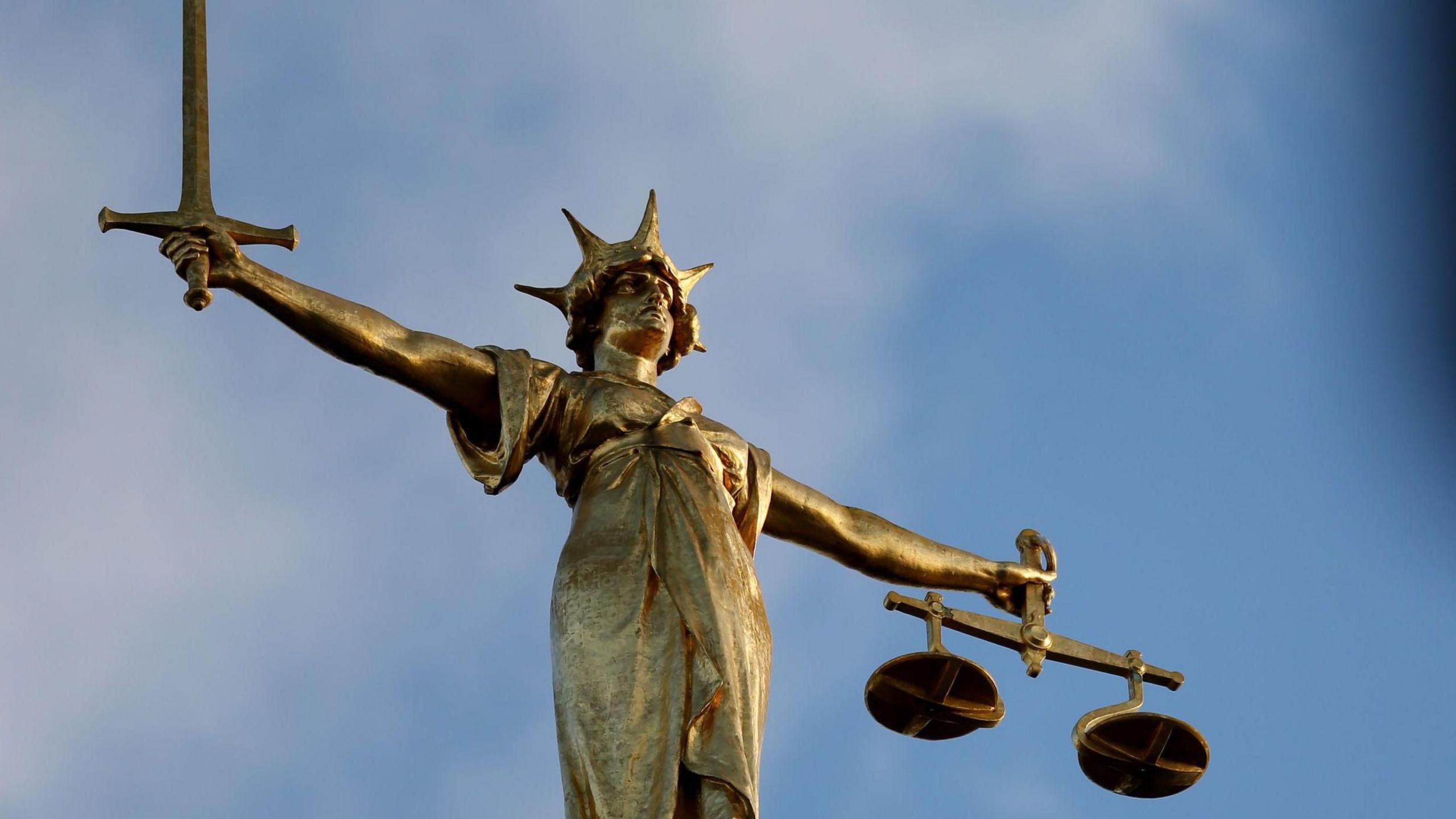 A golden statue symbolising justice. A female figure stands on a globe wearing a crown, holding a set of scales in one hand and a sword in the other.