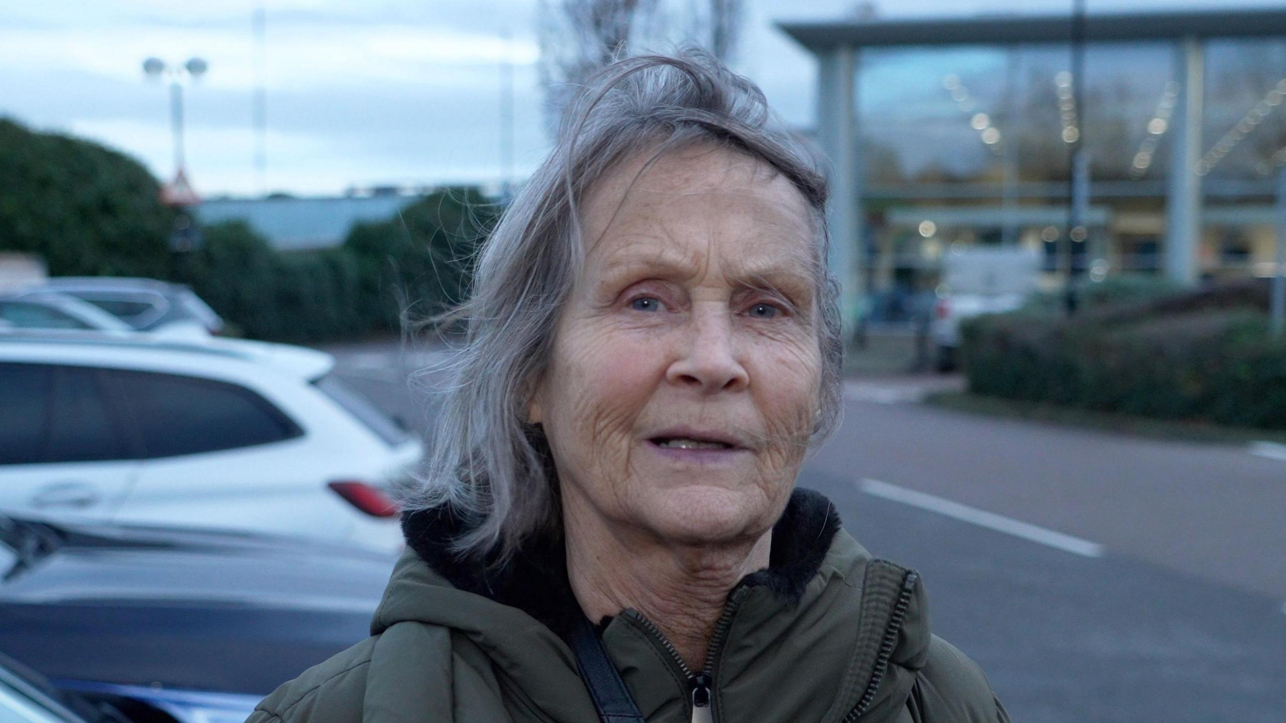 Pauline Strange standing next to some cars. She is wearing a green coat and has grey hair.