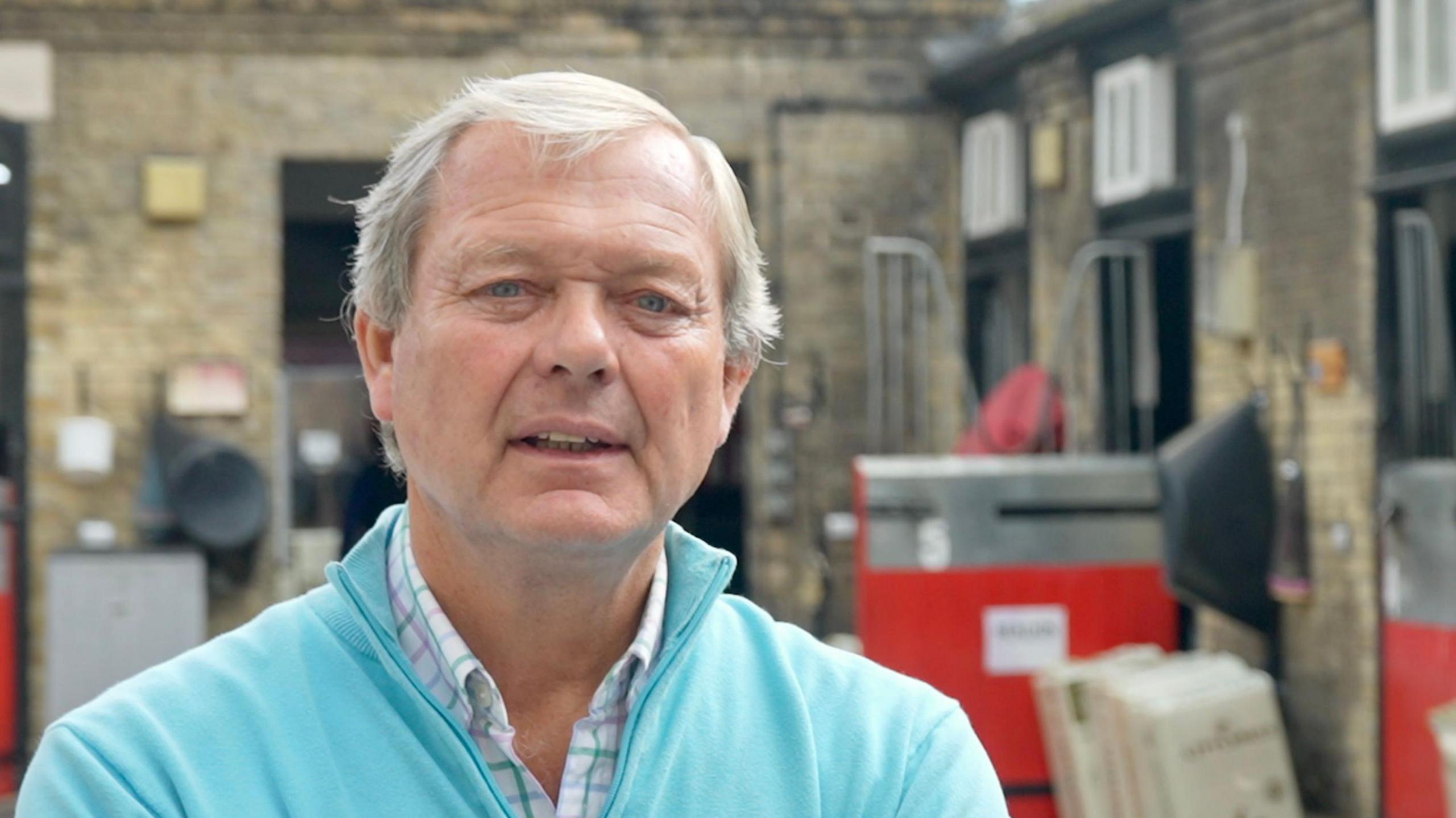 A head and shoulders shot of William Haggas. He is wearing a light blue jumper over a shirt. He is looking directly into the camera.