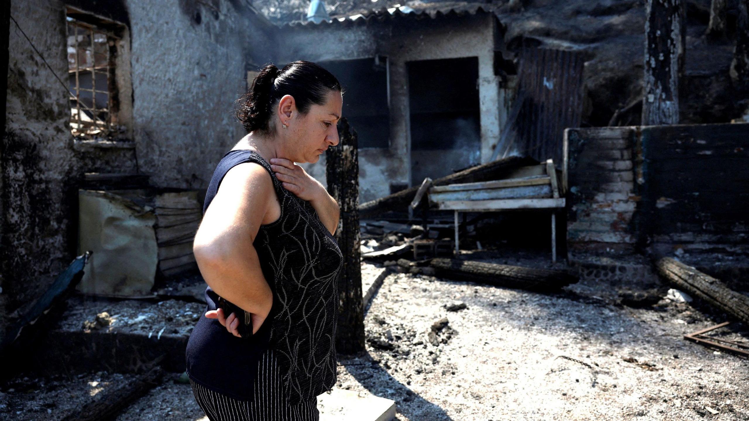 A lady with black hair wearing a black vest walks with her head down next to a blackened and charred burnt tavern