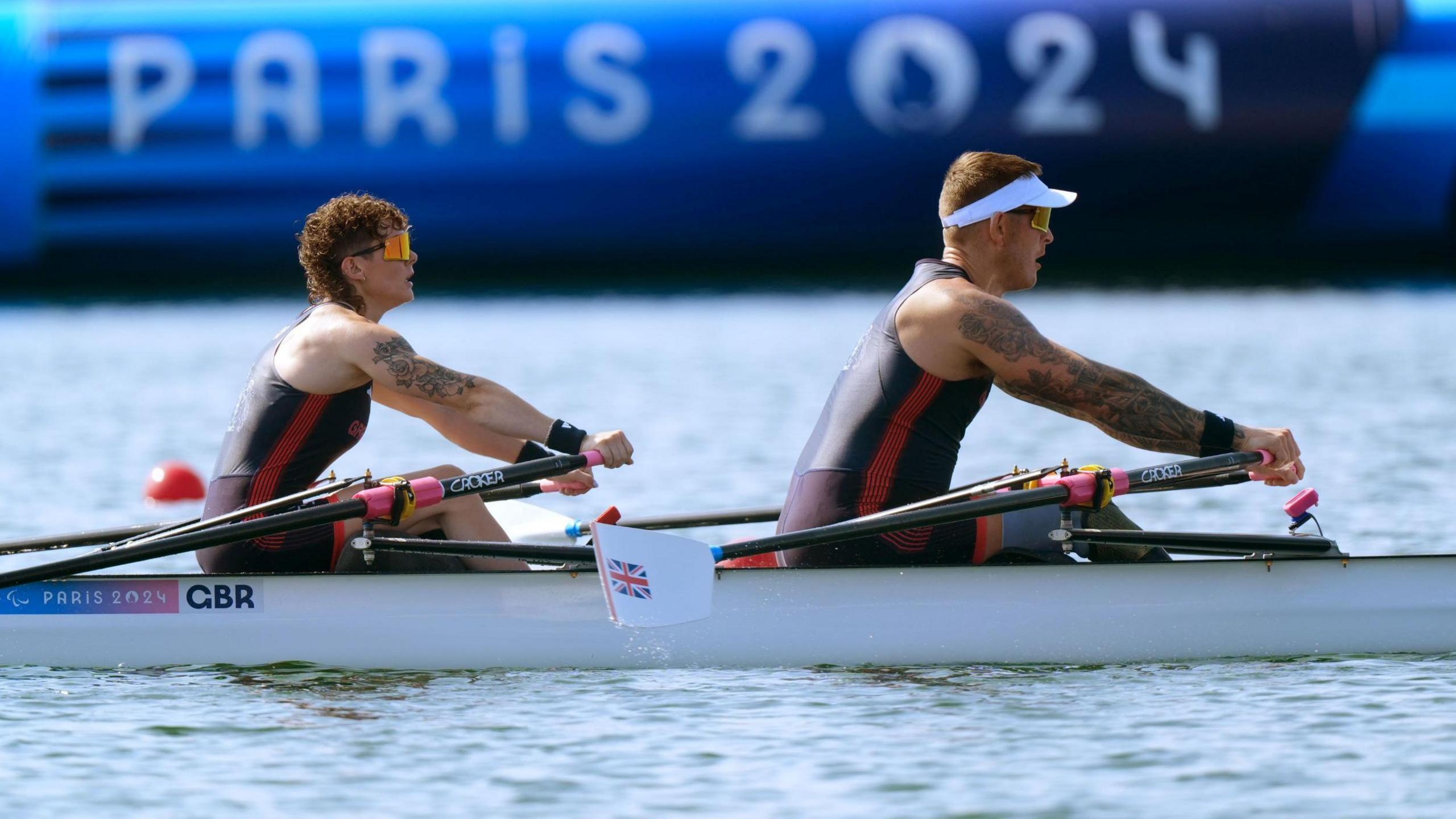 A man and a woman rowing a white boat. The woman sits at the back and has curly brown hair. The man sits at the front and has short blonde hair. They are both wearing sunglasses, and black and red wetsuits.