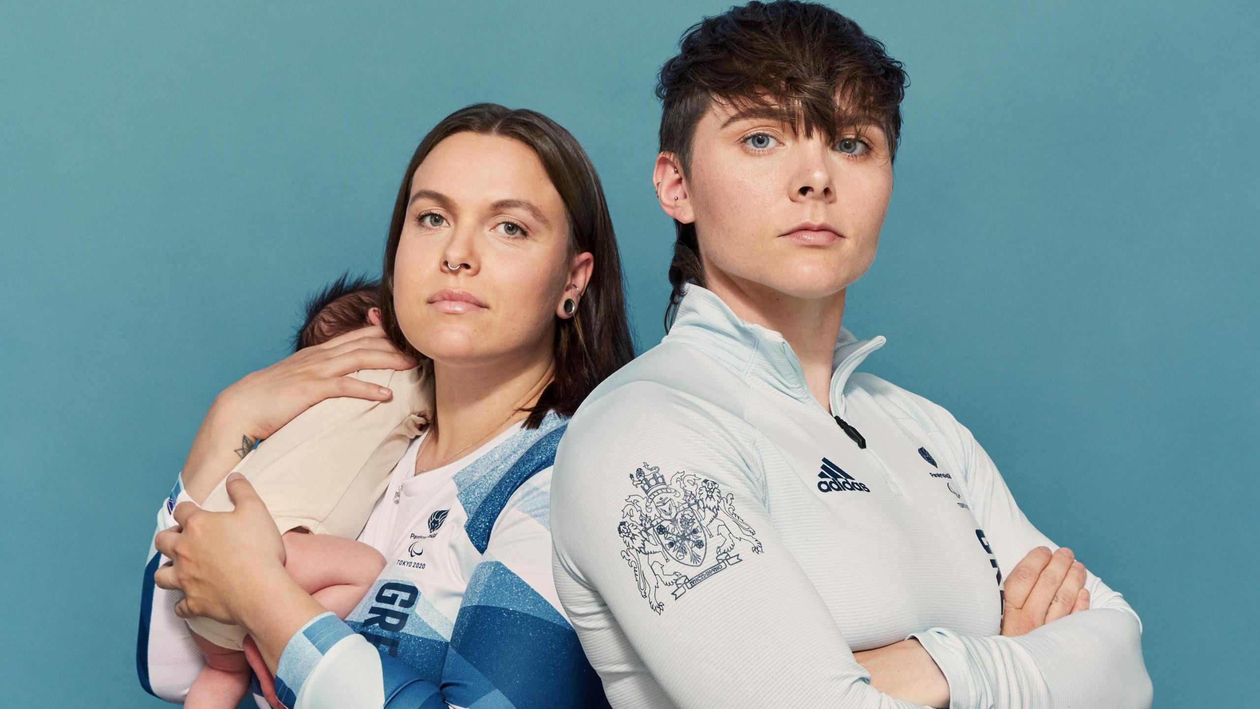 Two women stand side by side. The woman on the right has straight brown hair and she is holding a baby. The woman on the right has short brown hair. They are both wearing long-sleeved sports tops.