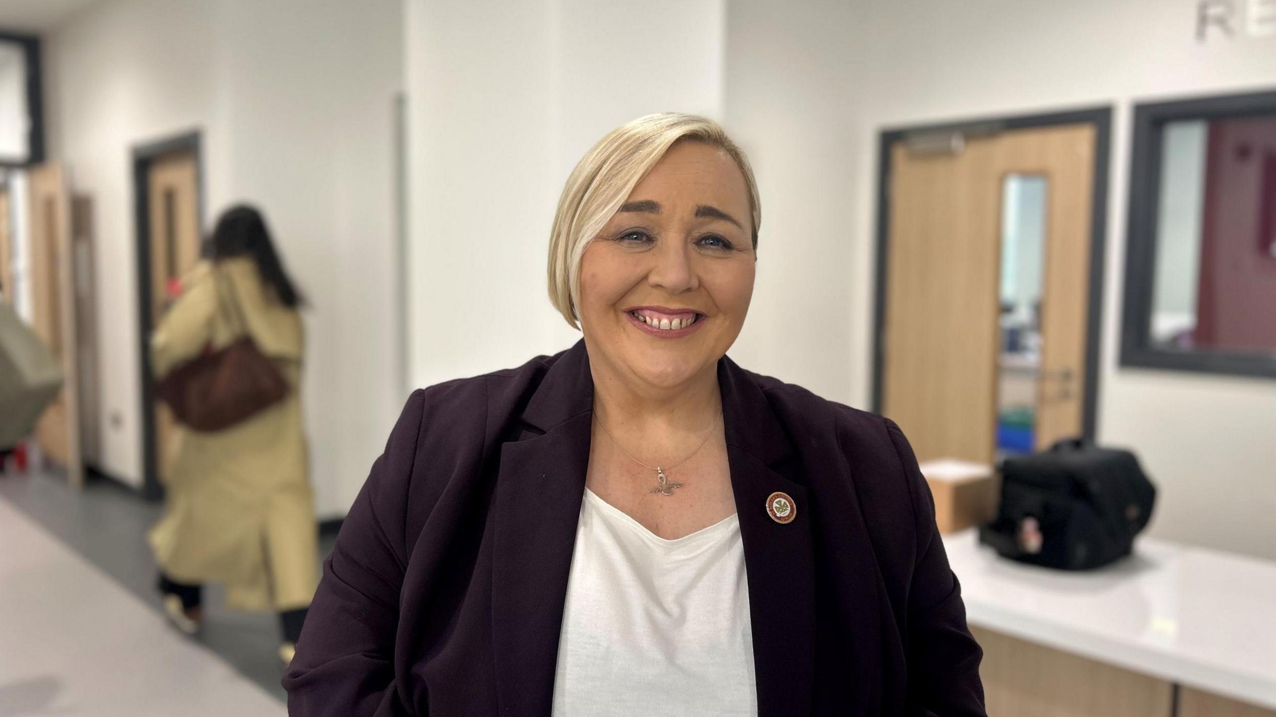 Fiona Kane is smiling widely at the camera. She has blonde, chin-length hair which she is wearing a side part. She is wearing a plum coloured blazer style jacket with a white t-shirt underneath. She is standing in a school corridor.