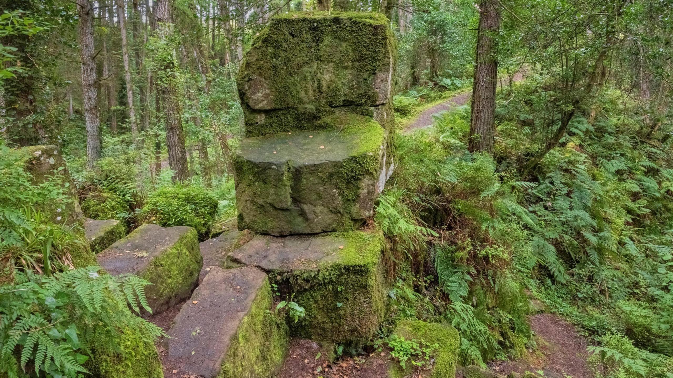 A large sandstone formation in the middle of some woods. The stone are piled on top of each other in the shape of a chair and are covered in moss.
