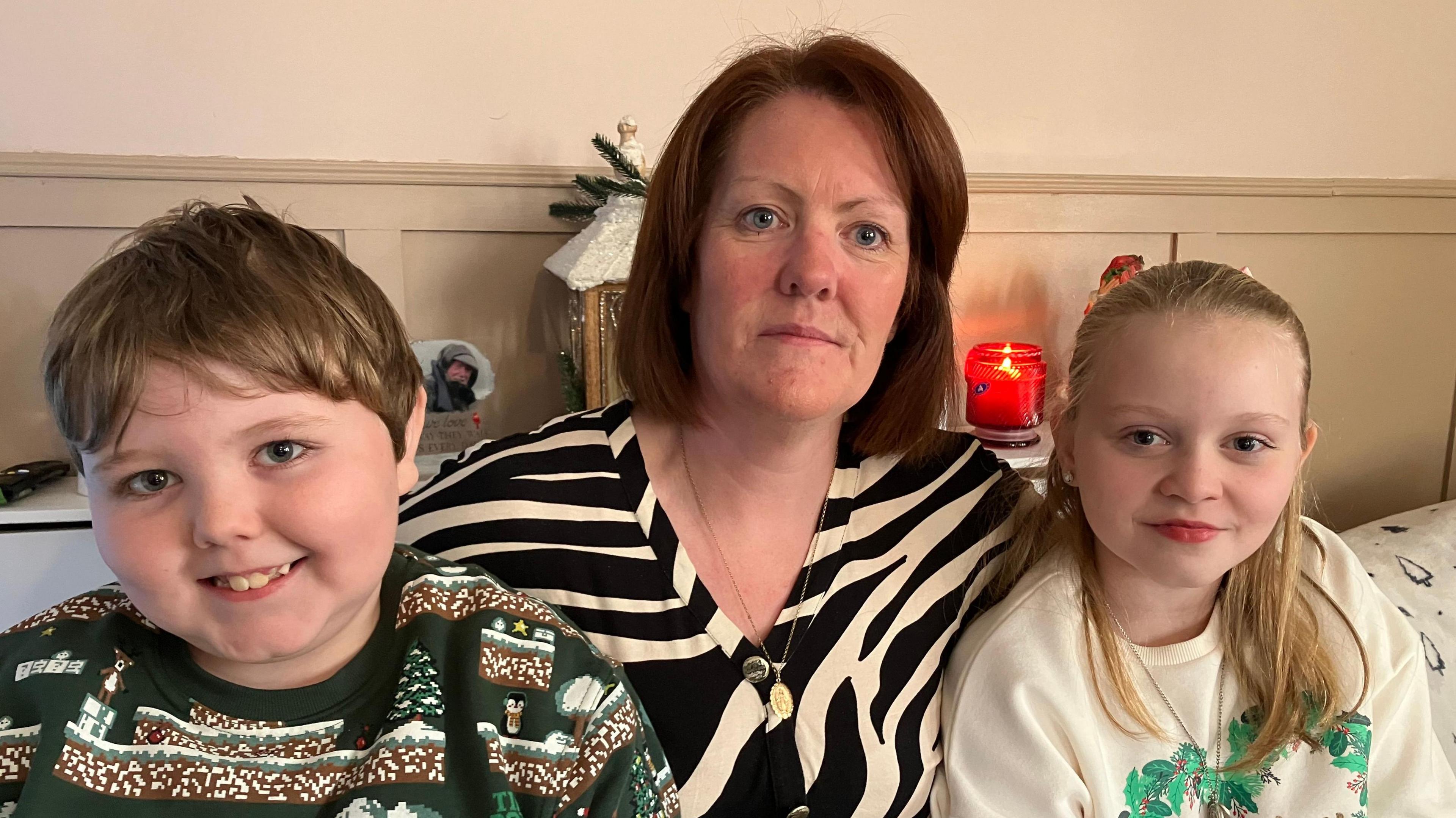 Shows a woman in a black and white zebra-patterned top with a young boy in a Christmas jumper to the left and girl with fair hair in light-coloured Christmas jumper to the right.