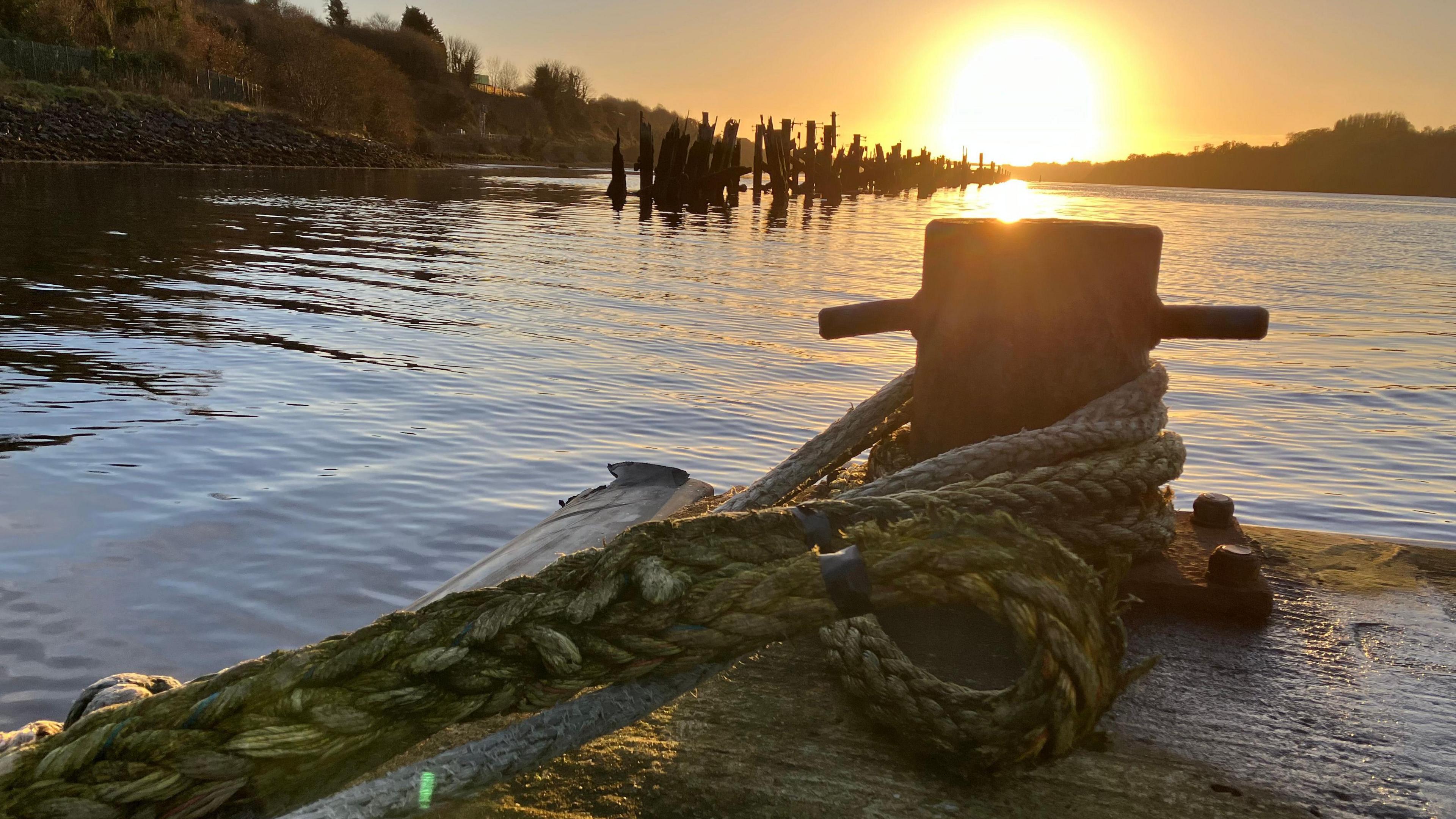 A thick mooring rope is coiled around a metal bollard at the corner of a pier. A bright sun is coming to rest on the horizon, its yellow centre fanning outward to a deep orange, before the sky, at the corners of the frame, becomes blue-grey. A crosshatch of ripples can be seen on the surface of the water and broken stilts of an old wharf jut from the water.