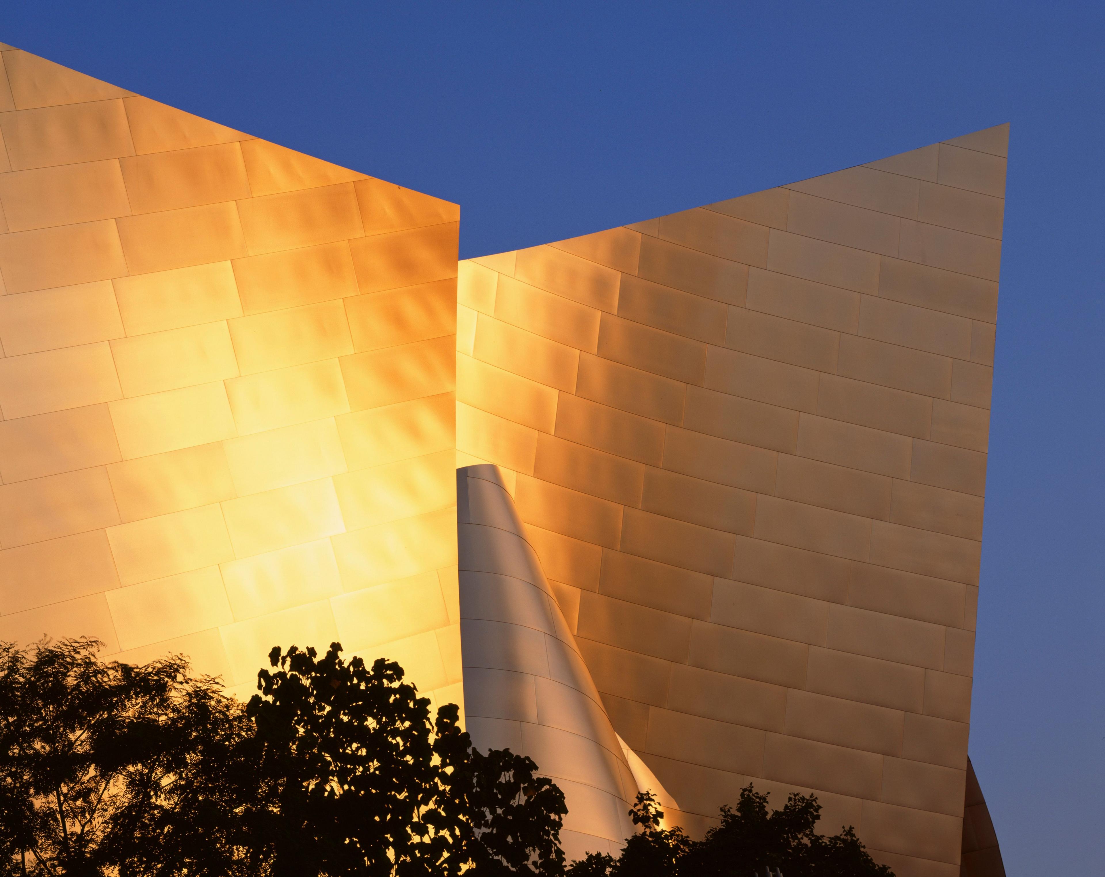 A golden hour view of the Walt Disney Concert Hall in Los Angeles. The sun reflects off the titanium cladding that is shaped like wings around a curved element. There are the outlines of trees in the foreground, with a blue sky framing the shot.