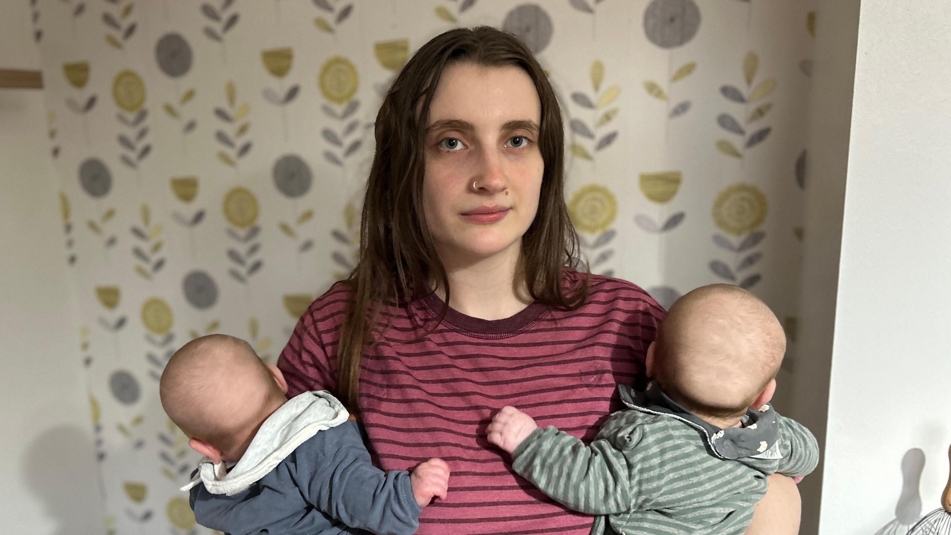 Woman with dark brown hair looking at the camera while holding two babies. She is wearing a red striped T-shirt and the babies are wearing navy and green.
