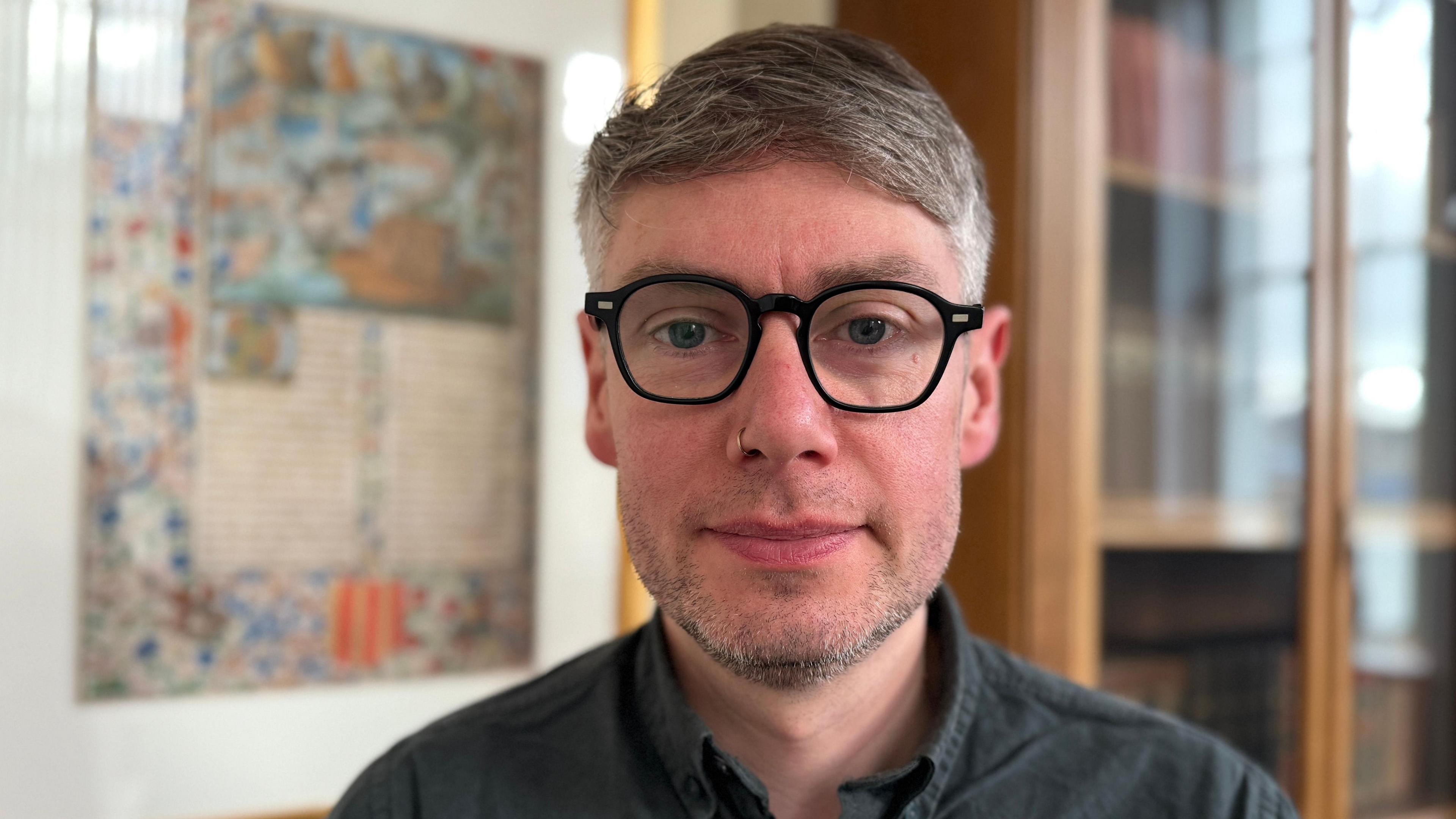 Chris Cassells, a man with short greying hair and glasses, standing in front of a bookcase and a wall chart.